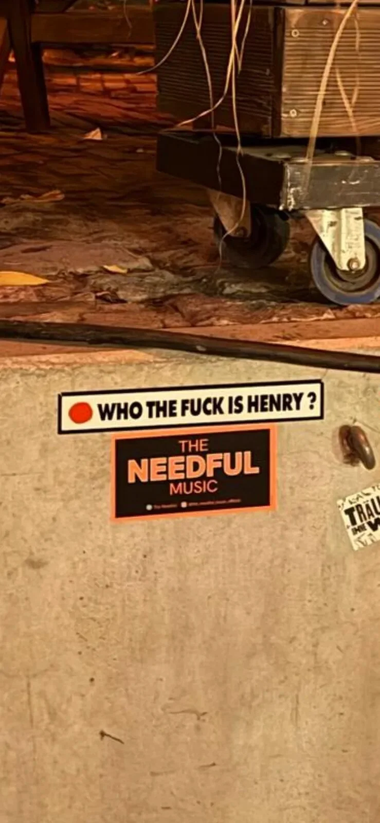 Close-up of a concrete surface with two stickers, one asking "WHO THE FUCK IS HENRY ?" and another for "THE NEEDFUL MUSIC." Part of a wooden structure or furniture on wheels is visible at the top, with some wires hanging down.