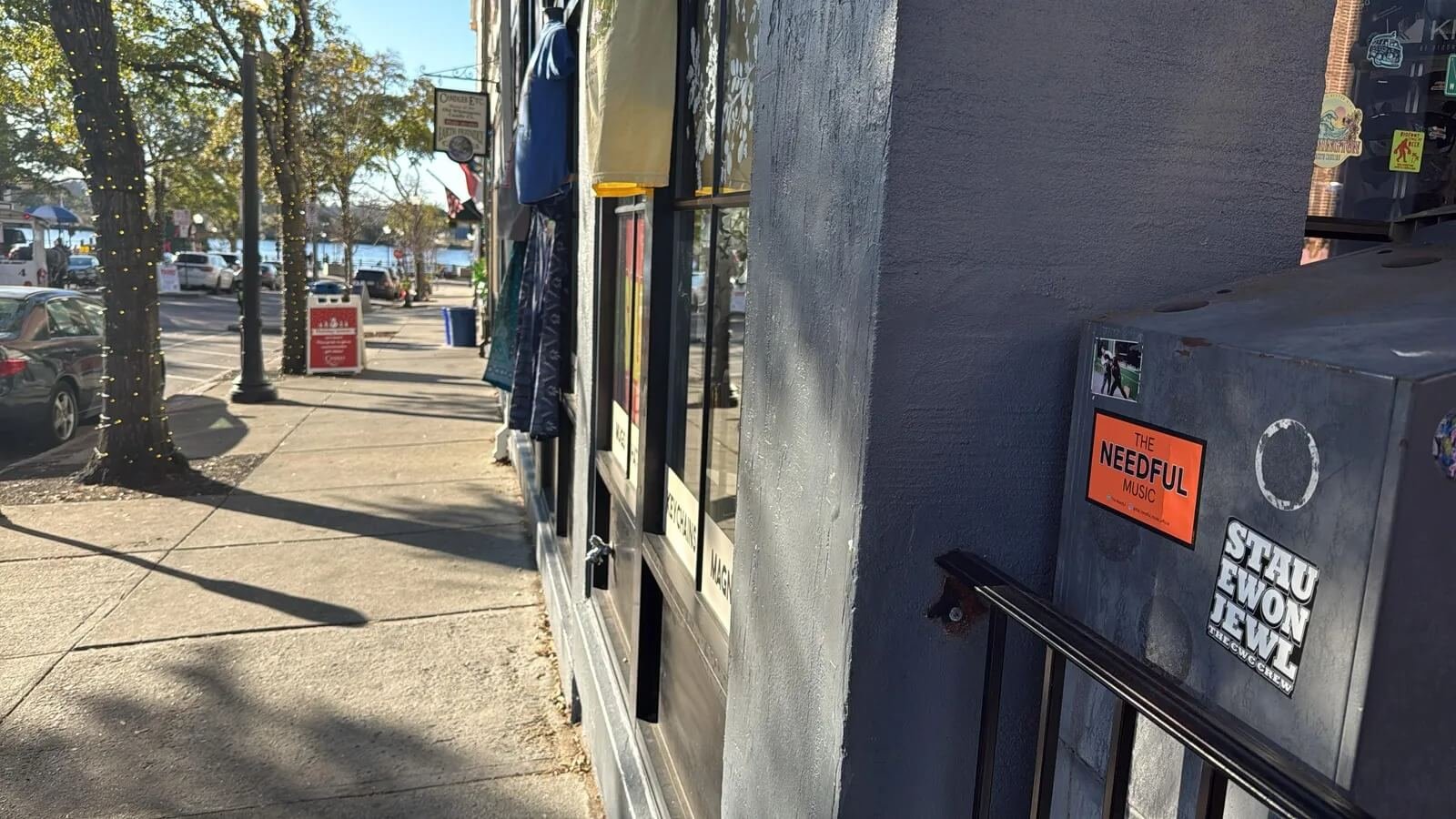 Sidewalk with parked cars, trees decorated with string lights, storefront with signs, and a utility box on the wall with stickers and an orange sticker reading 'The Needful Music'.