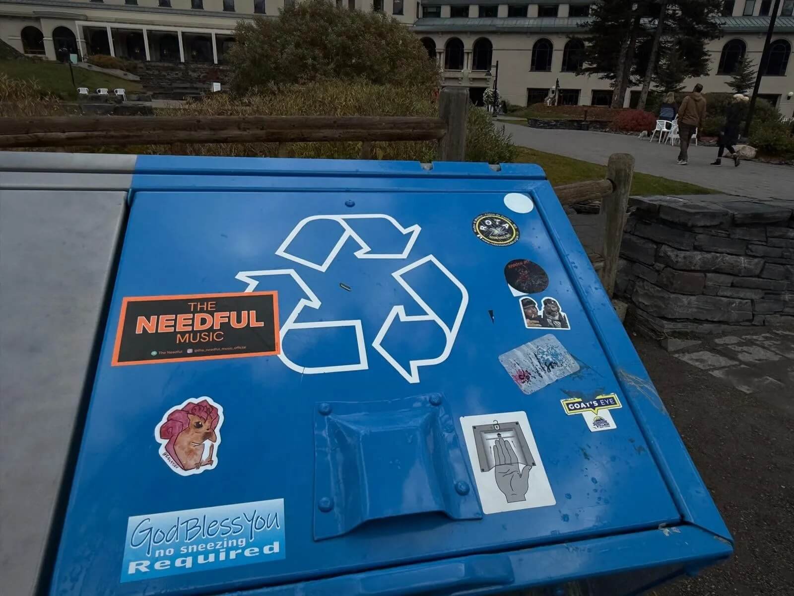 A blue recycling bin covered with various stickers, including for 'The Needful Music' located outdoors near a stone wall and a grassy area, with people walking in the background.