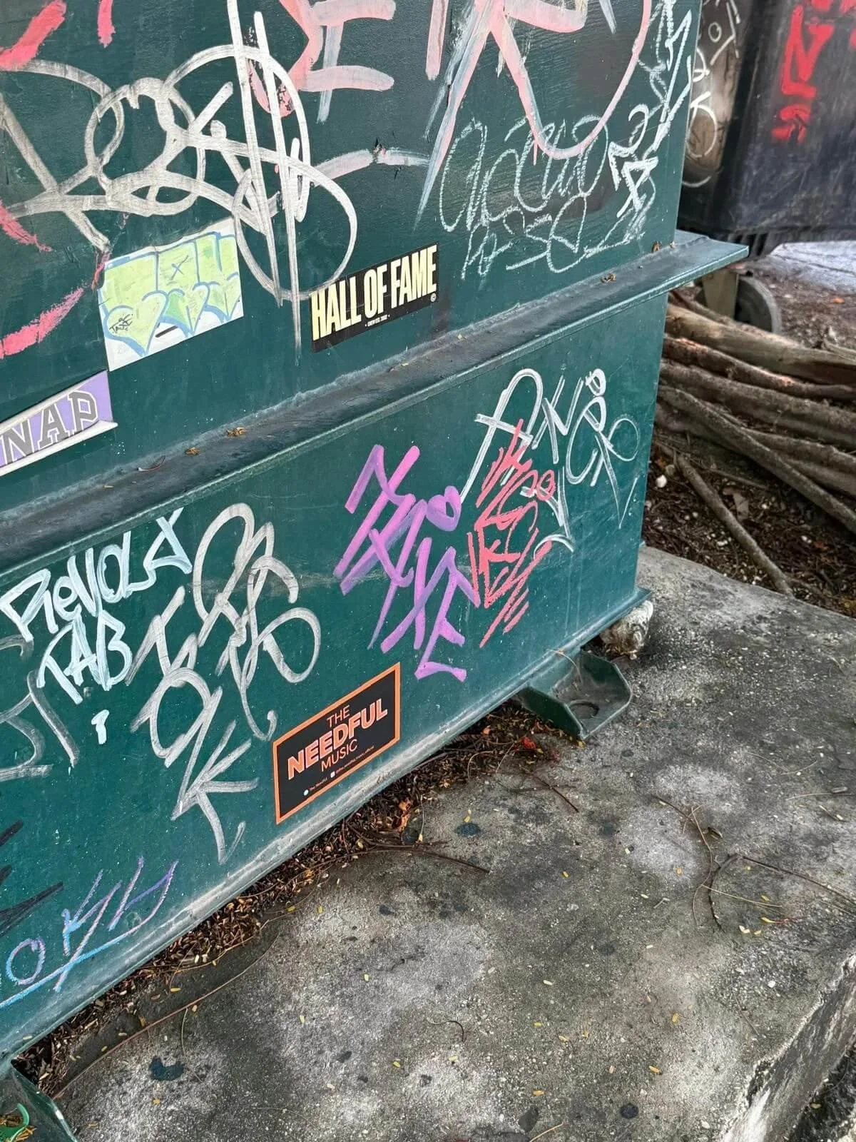 Green outdoor utility box covered with various colorful graffiti, stickers, and tags, located on a concrete surface with tree roots nearby.