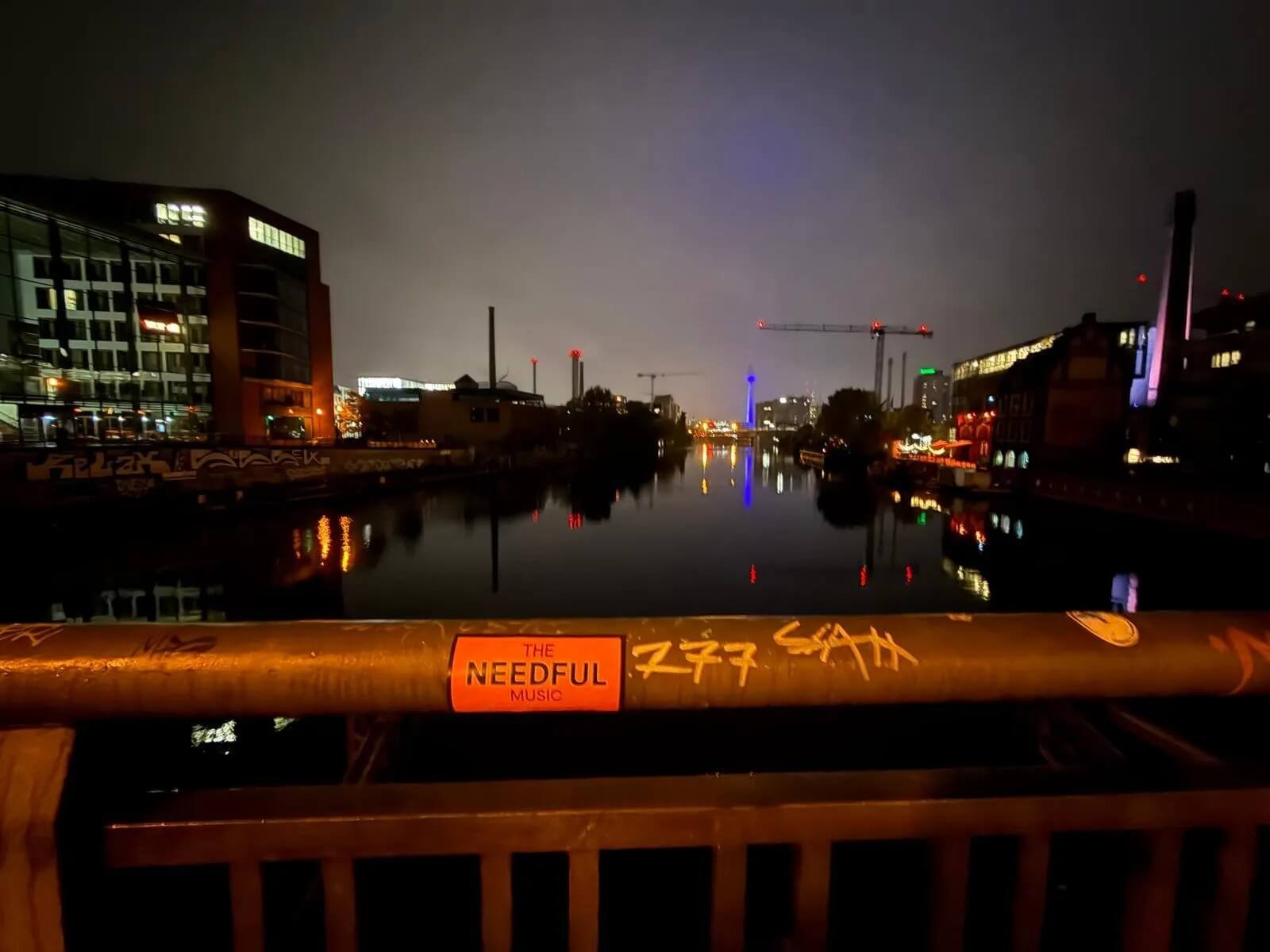 Night scene of buildings and a river reflecting city lights, with construction cranes and a bridge in the distance, seen from a bridge or walkway with graffiti and a sticker reading 'The Needful Music' in the foreground.