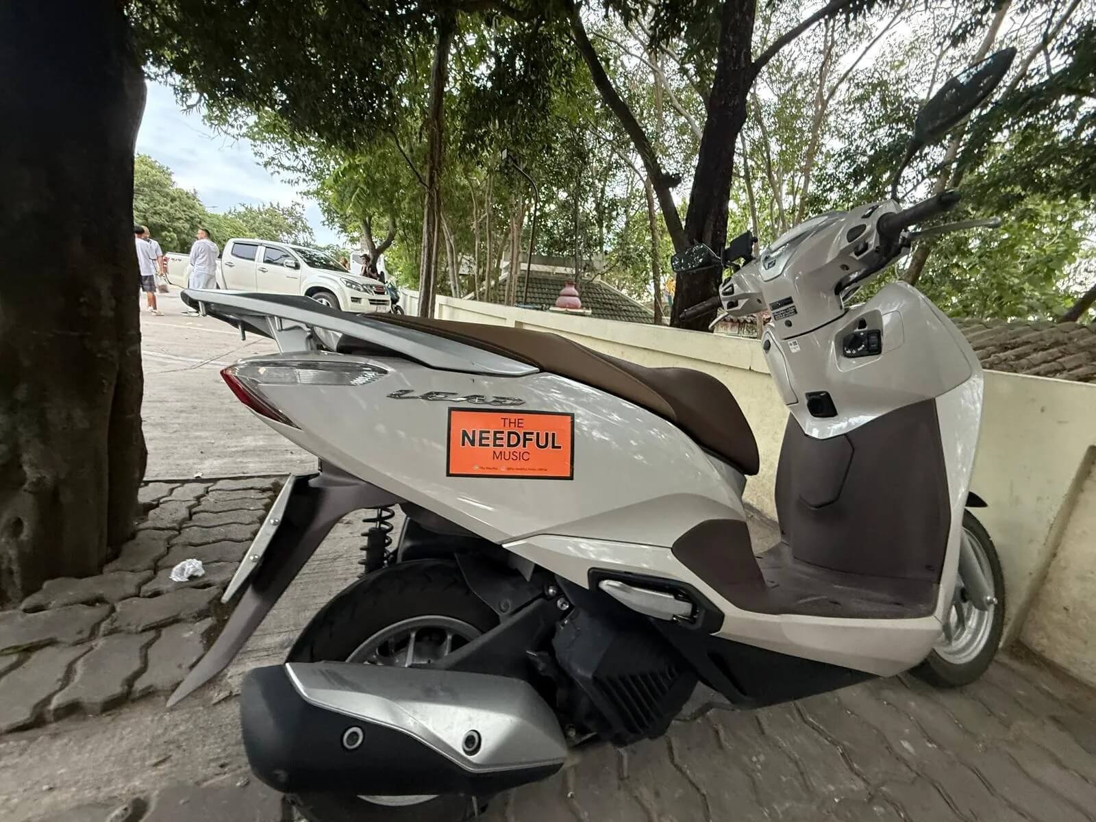 A parked white scooter with a brown seat, labeled "The Needful Music," on a cobblestone sidewalk next to a tree. In the background, there are parked white vehicles and a few people walking near trees and a building.