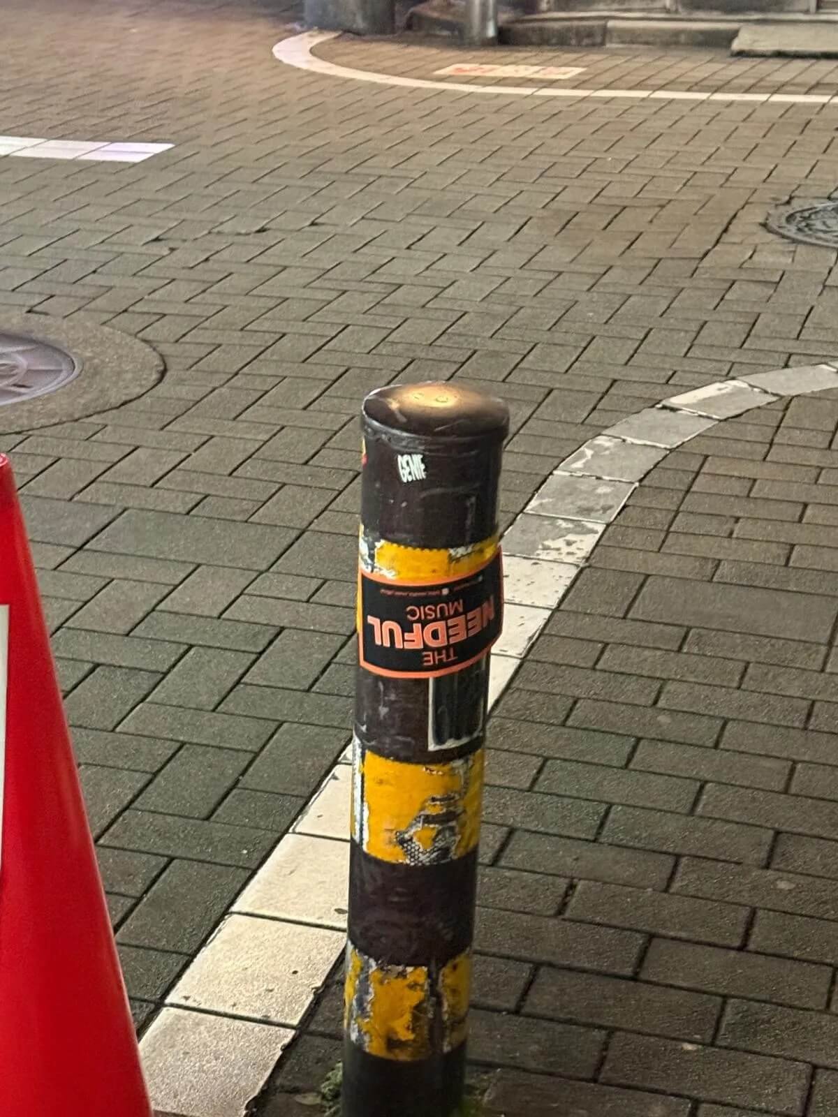 Black and yellow striped metal bollard with a sticker that reads 'The Needful Music' on a brick sidewalk at night.