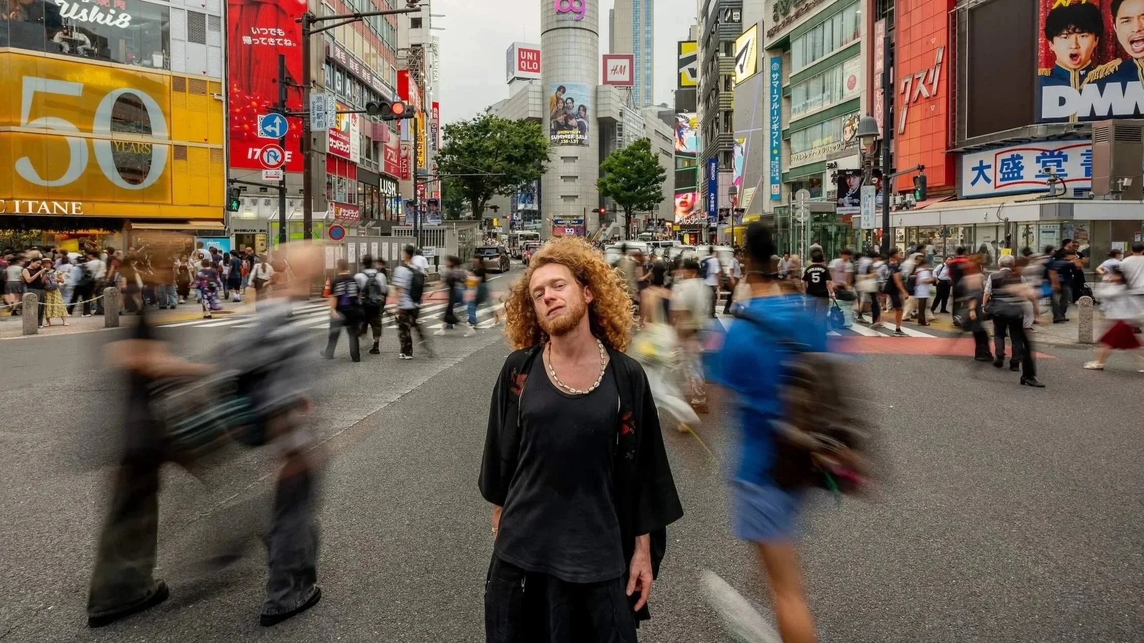 A man with curly red hair, wearing a black shirt and necklace, stands in the middle of a busy city street surrounded by a crowd of pedestrians and moving vehicles. The background features illuminated billboards and tall buildings, indicating a bustling downtown area.