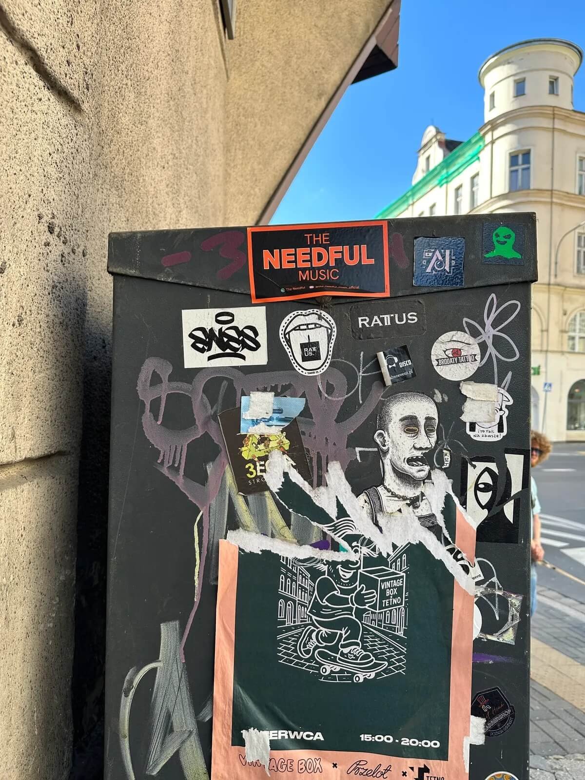Street-side poster board with various stickers and graffiti, including a black and white illustration of a person with a shaved head and tongue out wearing a headset, and a poster of a skateboarder. The board is leaning against a wall, with a beige b