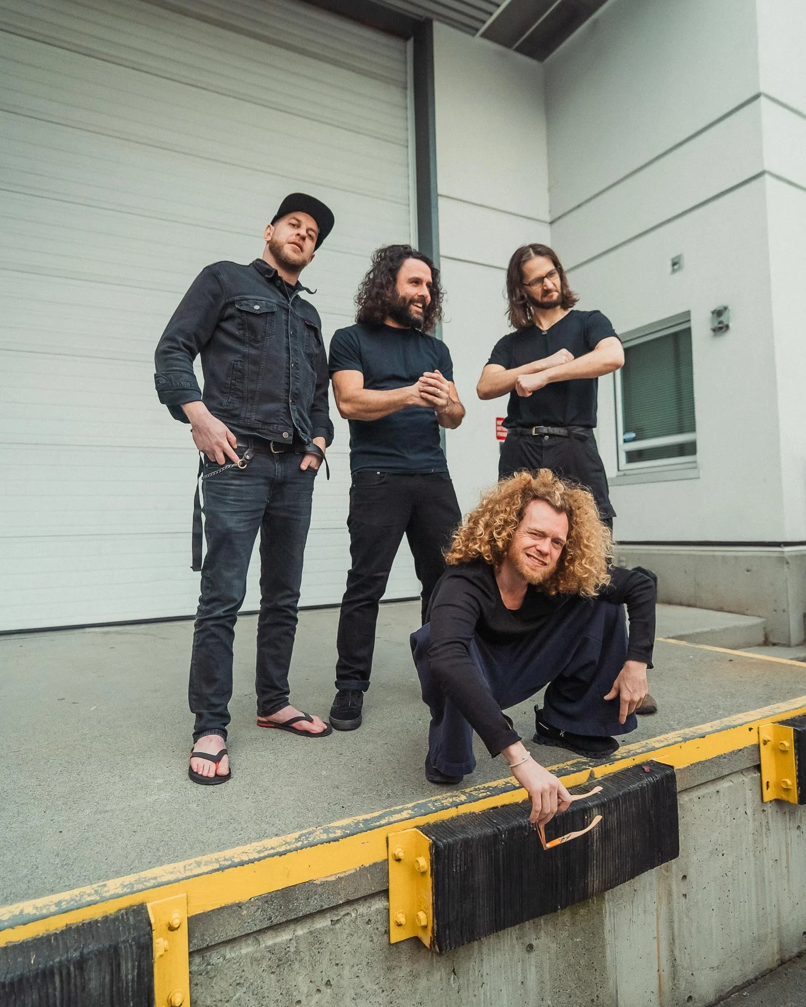 A group of four men, The Needful band, with one sitting on the ground and four standing behind a yellow and black safety barrier outside a building with a garage door, in casual black clothing.