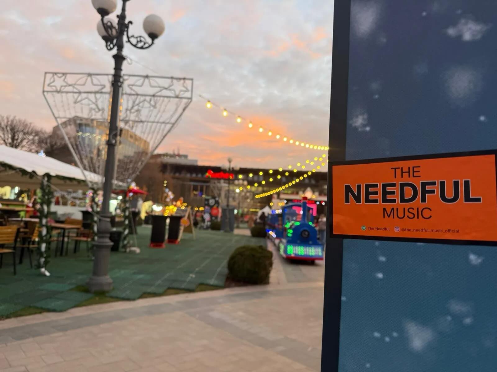 Outdoor market or fair at sunset with decorated tents, benches, a lighted train ride, and festive string lights in the sky. A sign in the foreground reads 'The Needful Music'.