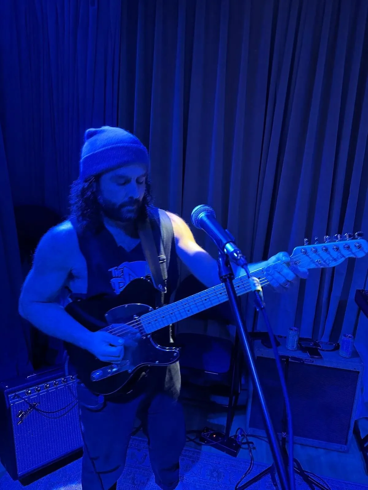 Kurt of The Needful with long curly hair, a beard, wearing a beanie, playing an electric guitar and singing into a microphone in a dimly lit room with blue lighting and wooden paneling in the background.