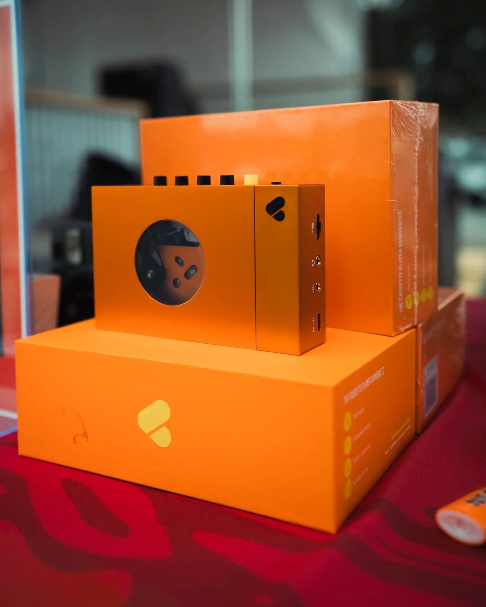 Close-up image of orange bluetooth 80's style walkman in packaging, with the box partially visible, set on a reddish surface.