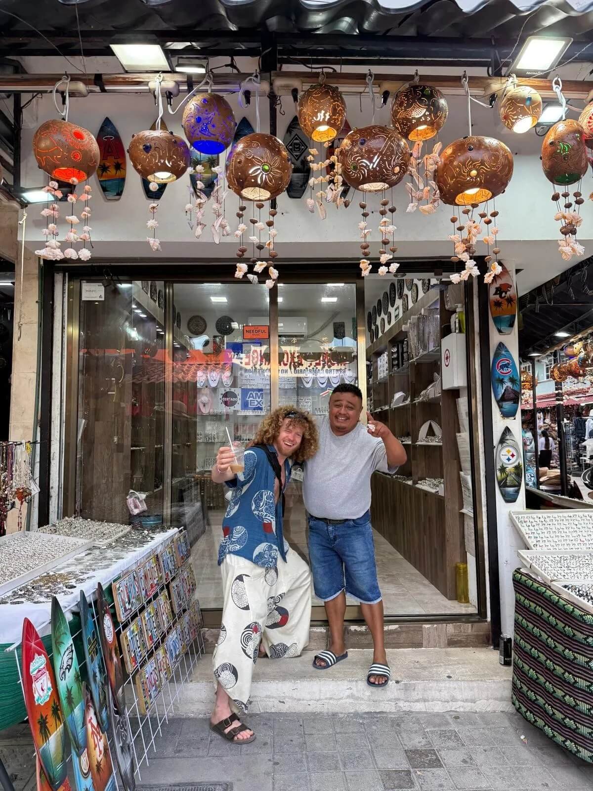 Glenn and another man standing outside a souvenir shop with hanging lanterns above. One man drinks a beverage, the other points at the camera, both smiling.