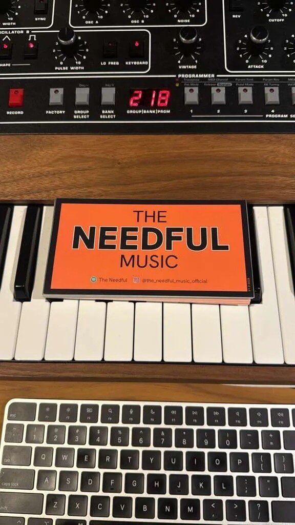 A close-up view of a keyboard, a digital music device, and a sign that reads 'The Needful Music' resting on piano keys.
