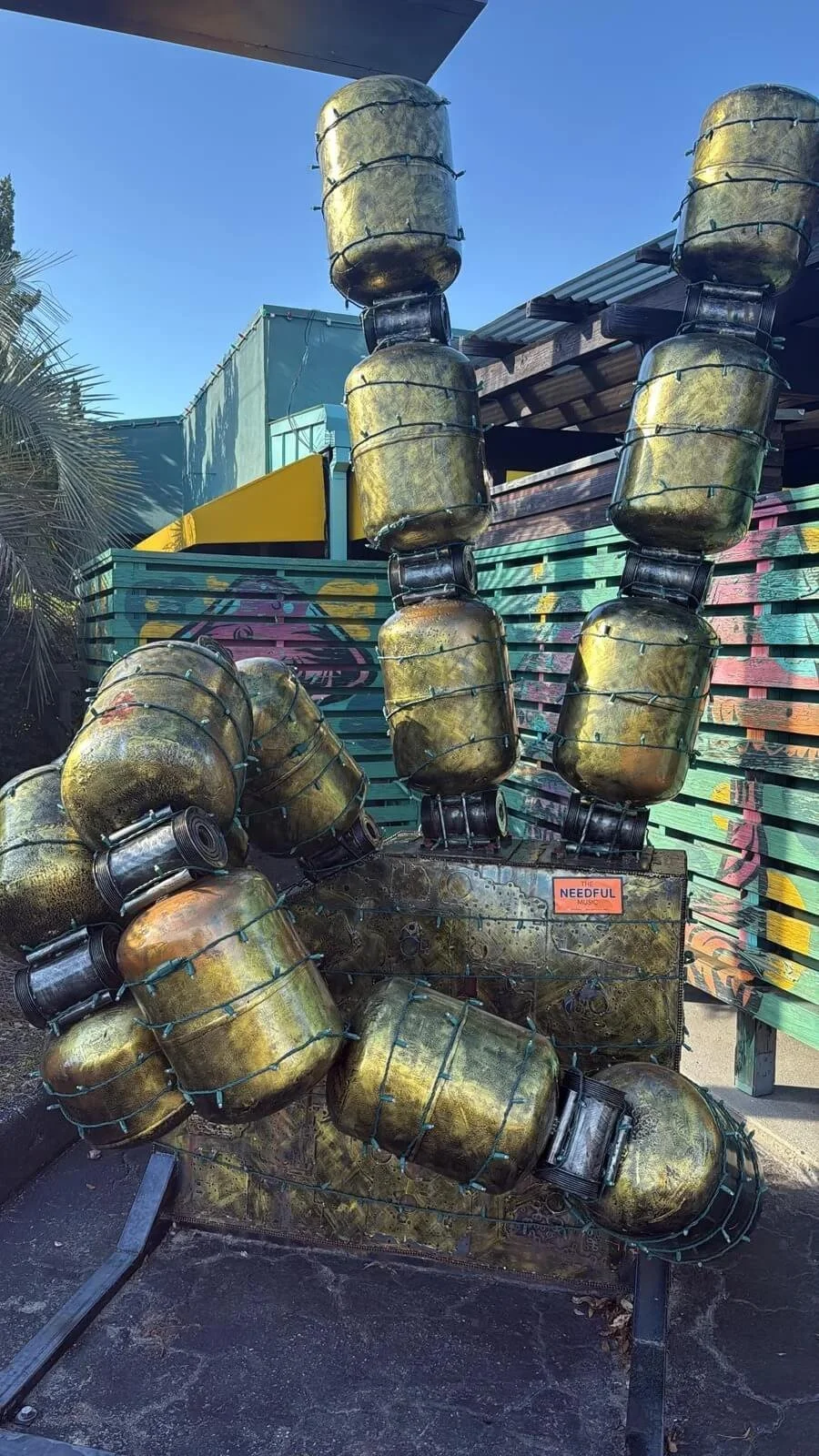 A large outdoor sculpture made of metal containers resembling a festive arrangement. The containers are painted to look like gold and are connected with wire, arranged in a way that resembles a palm tree or plants with a sticker entitled 'The Needful