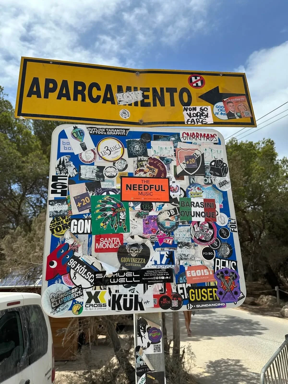A street sign in Italy with a yellow background and black lettering that reads "Aparcamento" (Parking) with a right arrow. The sign is covered with numerous stickers, some with images, text, and logos, including one reading "The Needful Music" and ot
