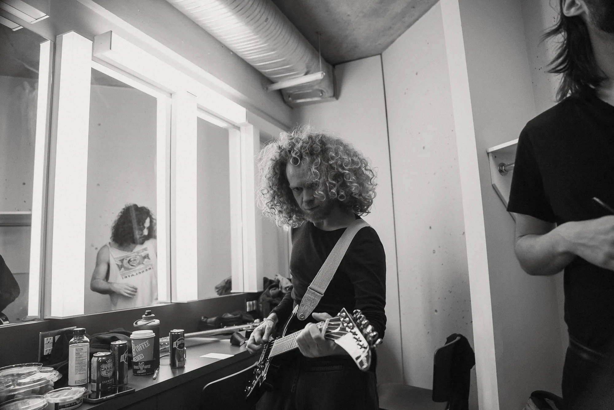 A person with curly hair playing an electric guitar in a dressing room, with a mirror and various items on the counter, including coffee cups and cans, and two other people partially visible.