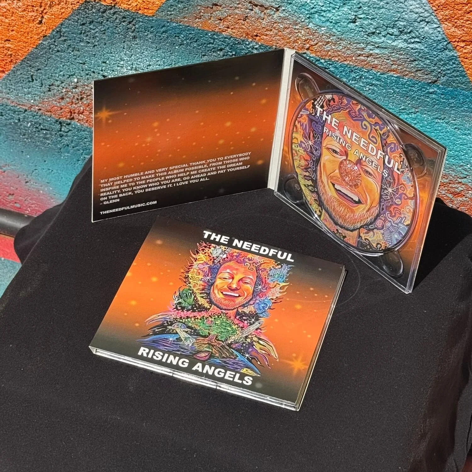 CD case and booklet with colorful psychedelic artwork, titled 'Rising Angels' by The Needful, placed on a black surface with a multicolored textured background.