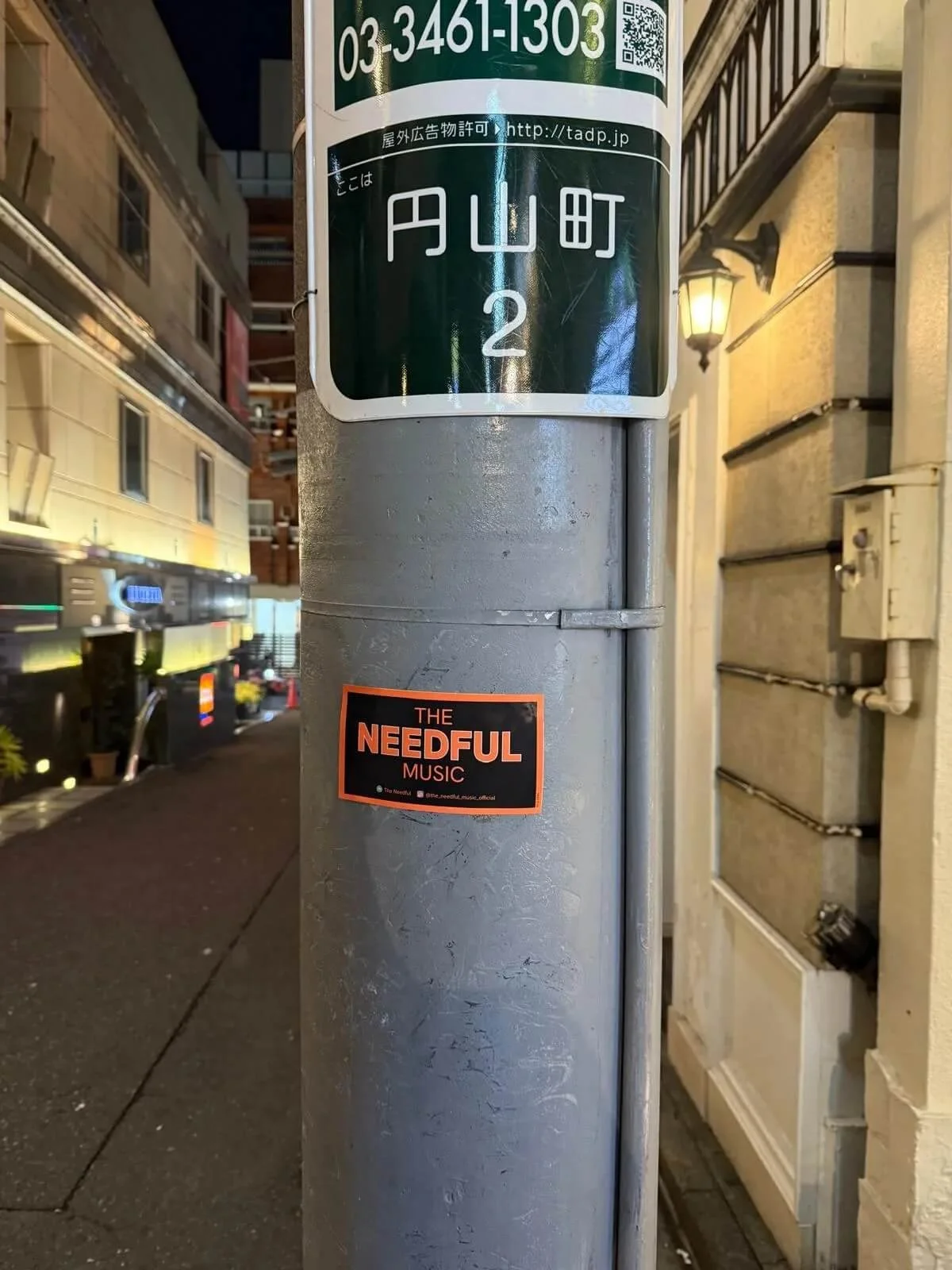 Street sign with Japanese characters, street number 2, and a sticker reading 'The Needful Music' on a gray pole at night.