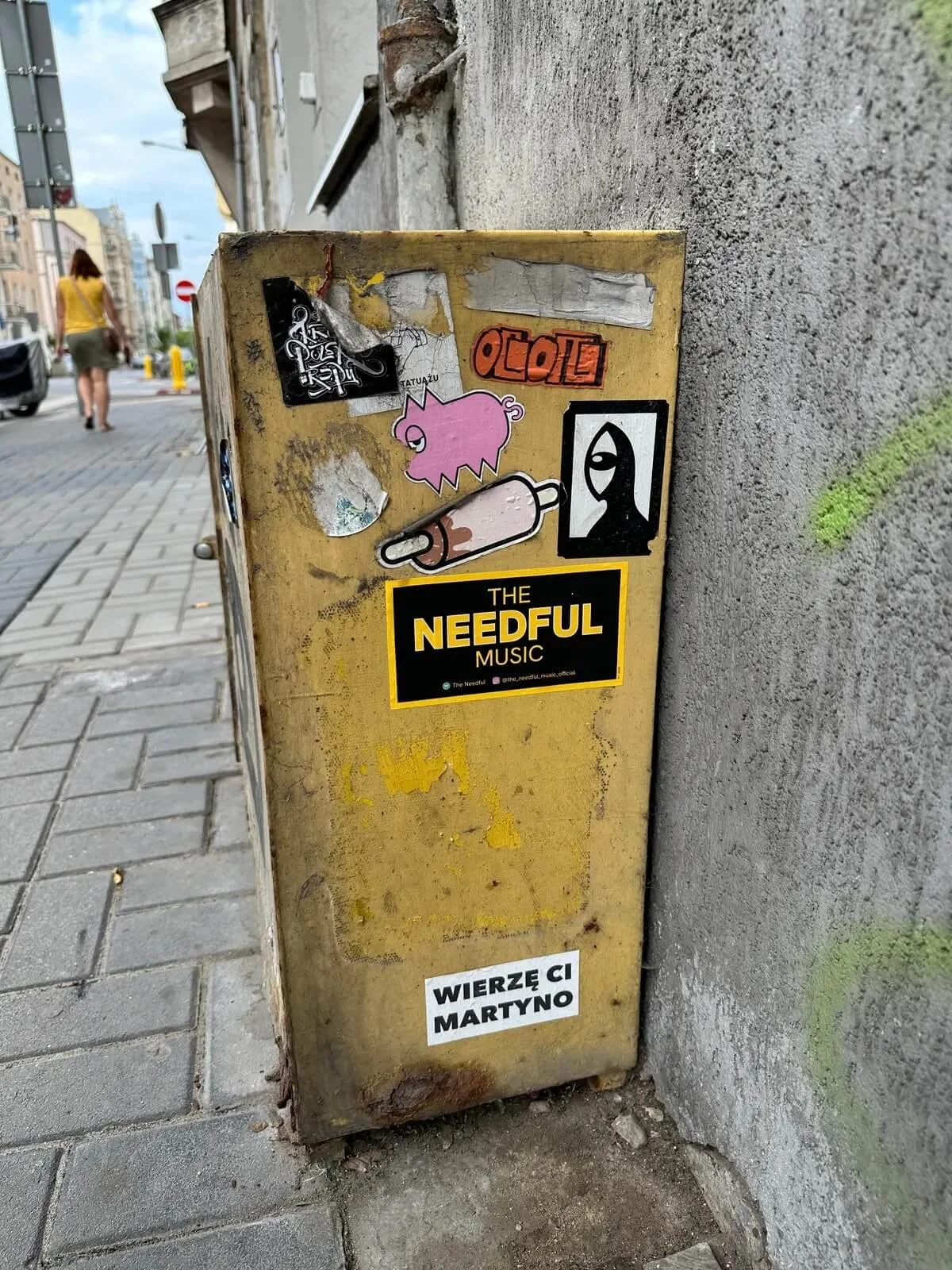 A weathered yellow electrical box on a city sidewalk, covered with multiple stickers, including a pink pig, a music band sticker for The Needful Music, and other graffiti stickers; a person walking, parked cars, and buildings are visible in the backg