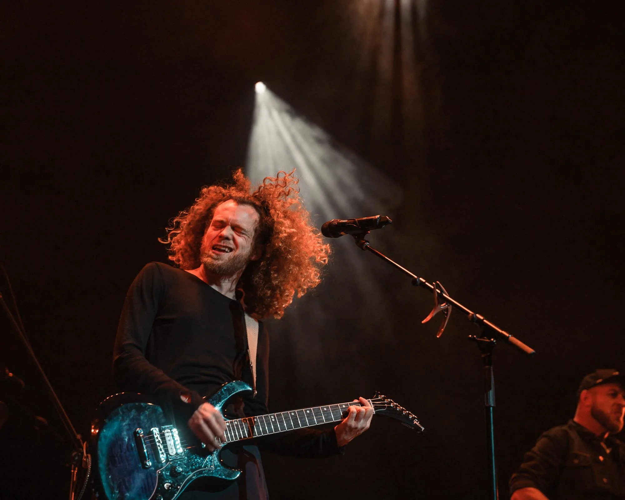 Glenn with long curly hair and a beard is performing on stage, playing an electric guitar and singing with an emotional expression. A spotlight shines down on him against a dark background.