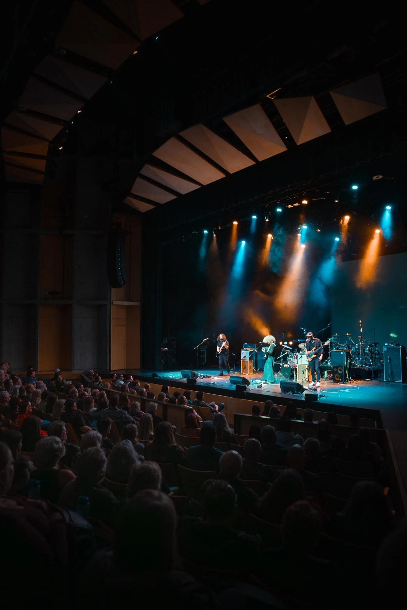 A live performance of The Needful on stage with colourful lighting, while an audience sits in a theatre watching.