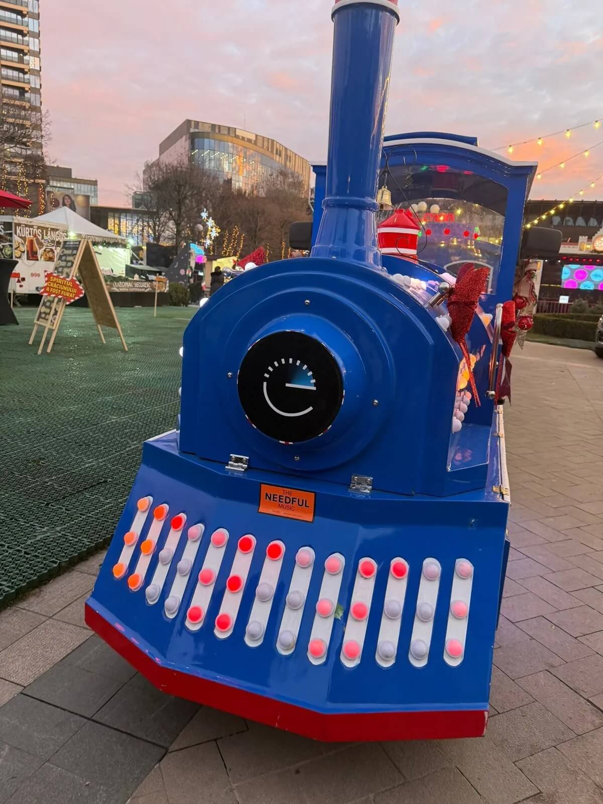 A blue train-shaped candy or snack vendor cart at an outdoor holiday market, decorated with red bows and string lights, with modern buildings and market stalls in the background at sunset.
