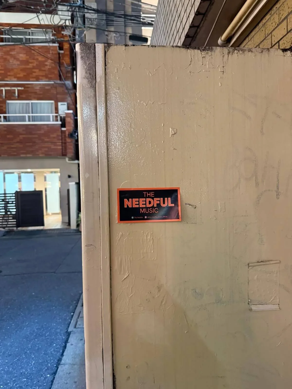 A small rectangular sticker on a beige wall with the text 'The Needful Music' in black and orange letters.