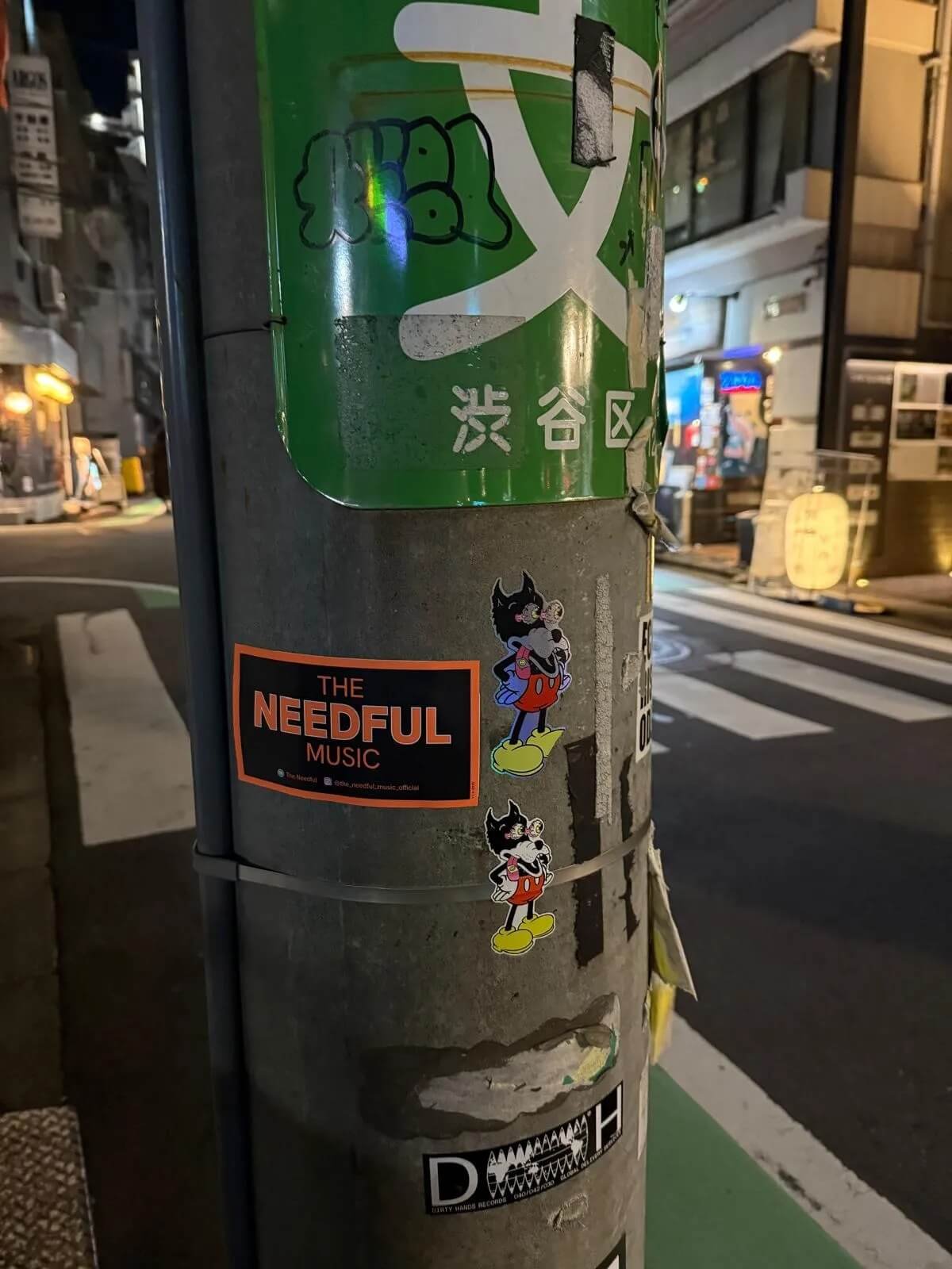 Street pole with multiple stickers, including a green sign with white Japanese characters, a black and orange 'The Needful Music' sticker, and cartoon character stickers of a black and white dog with yellow shoes.