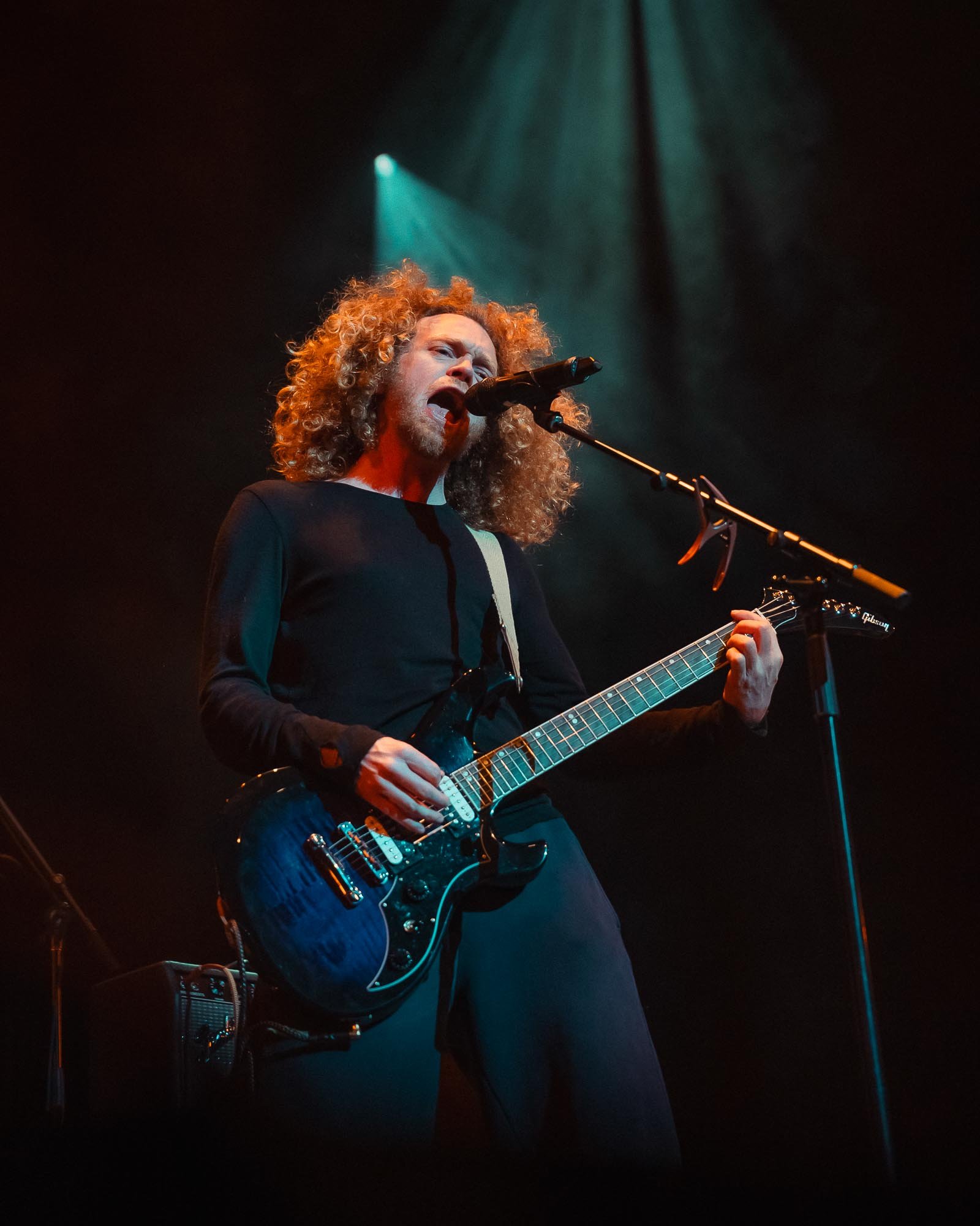 A male musician with curly hair passionately singing into a microphone and playing an electric guitar on stage, with stage lights shining behind him.