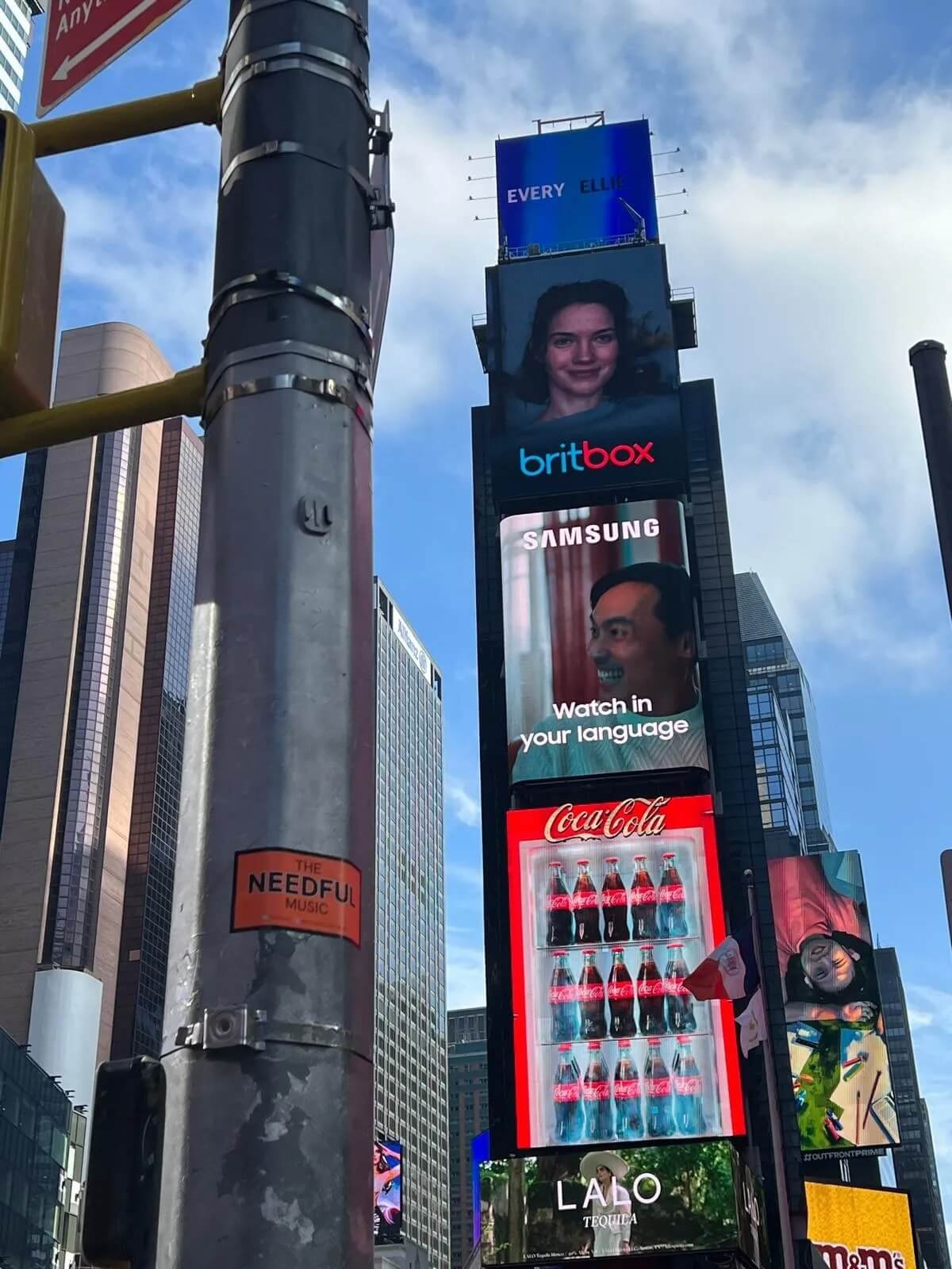 Multiple electronic billboards displaying advertisements in a cityscape, including a woman's face, text 'britbox', a man's face with the text 'Samsung Watch in your language', Coca-Cola bottles, and additional ads at the bottom, with a clear blue sky