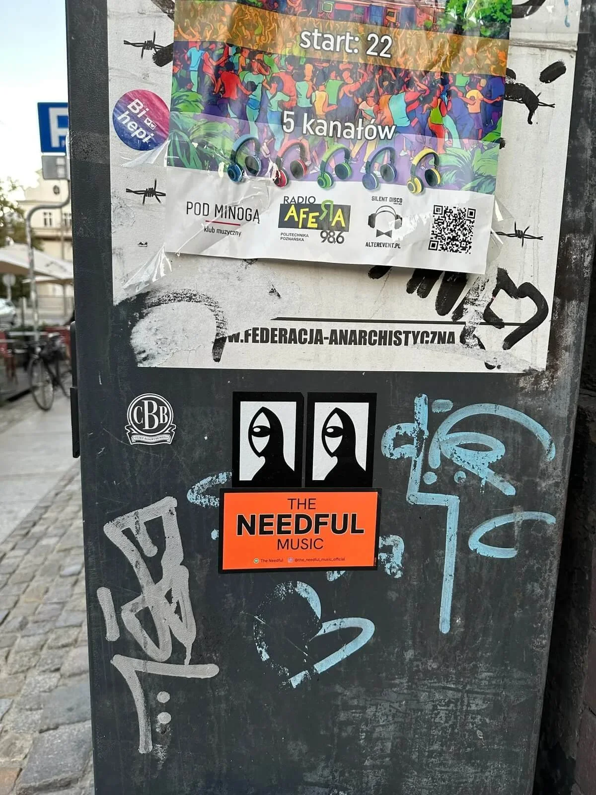 A black metal signboard covered with stickers and posters. There is a colorful poster at the top advertising an event with five headphone icons and the start time 22. Below, there is a sticker for the anarchist federation, and a sticker for the band 