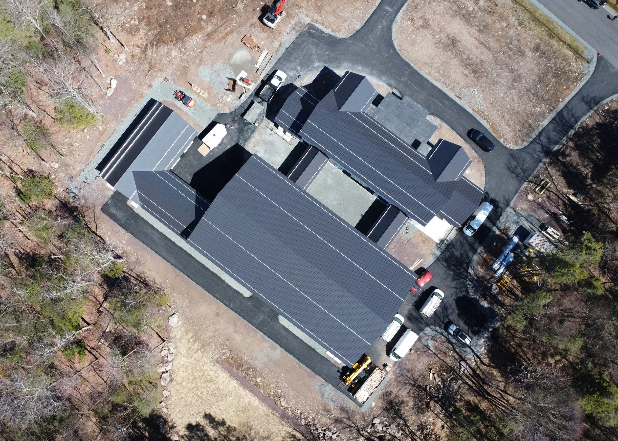 An aerial view of a newly constructed building with metal roofs, surrounded by parked vehicles, trees, and construction materials.