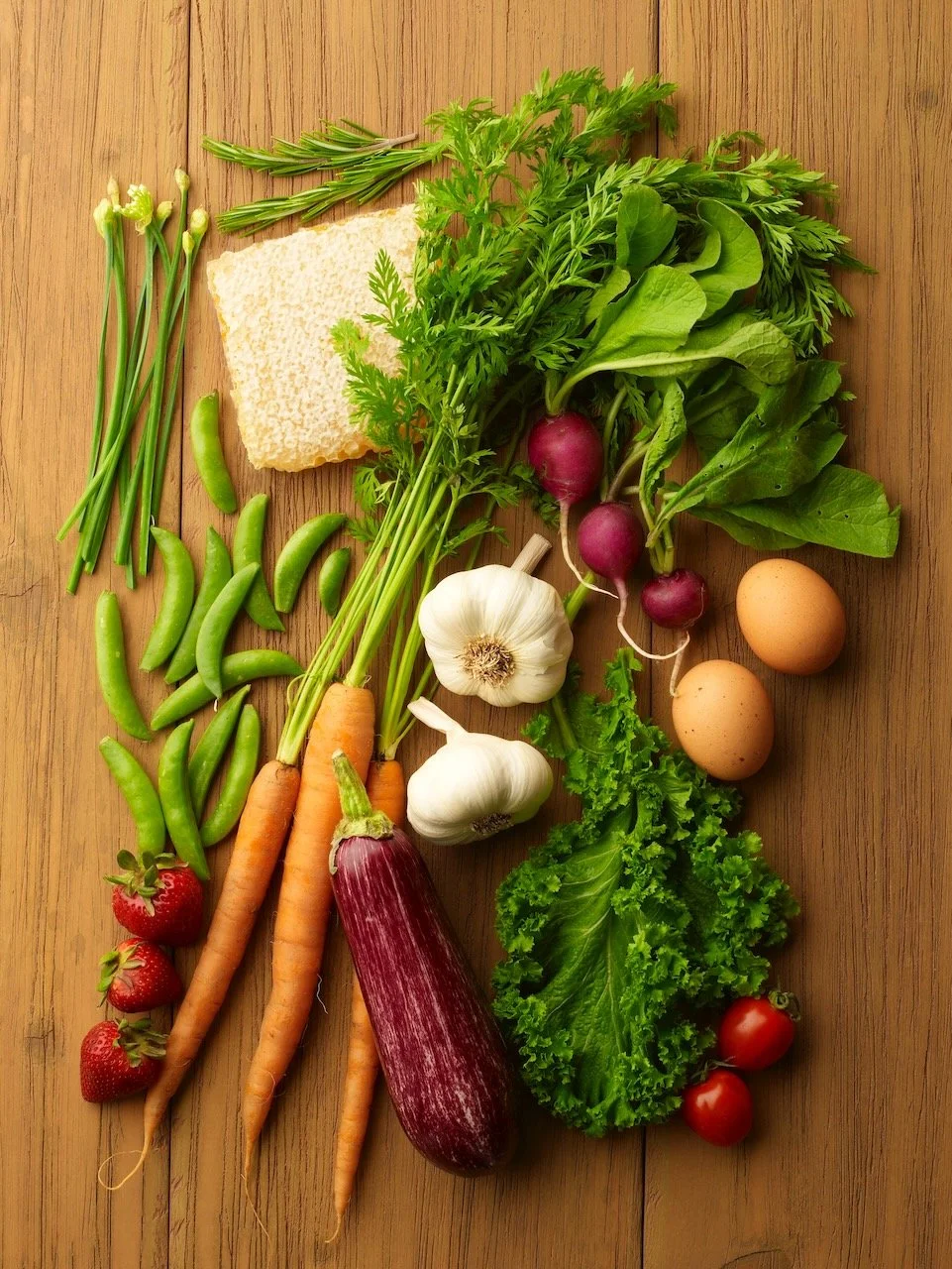 Fresh vegetables and herbs including strawberries, carrots, garlic, eggplant, lettuce, tomatoes, radishes, eggs, beans, herbs, and a piece of sponge cake on a wooden surface.