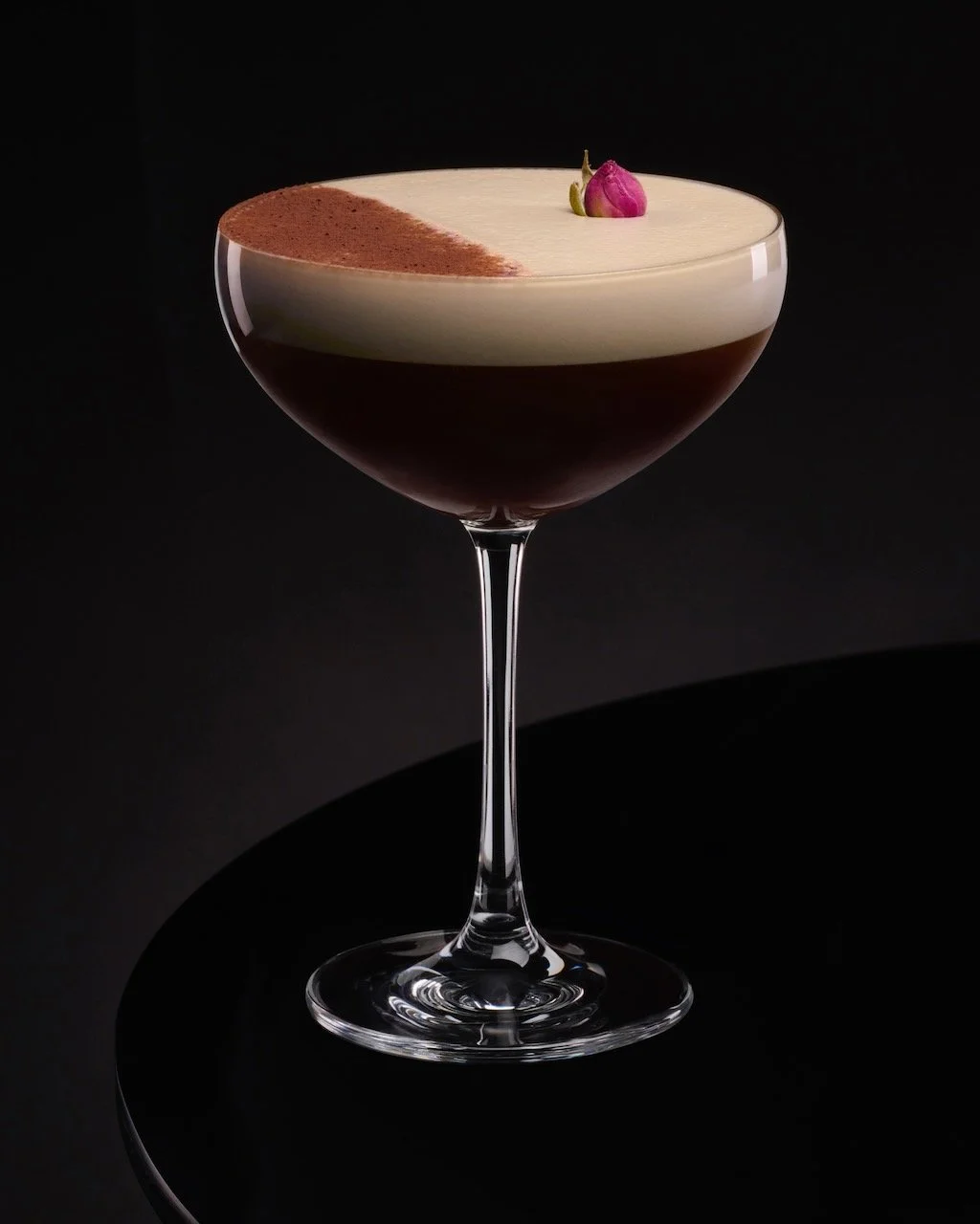 A cocktail in a coupe glass with a layered appearance, garnished with a small pink flower.