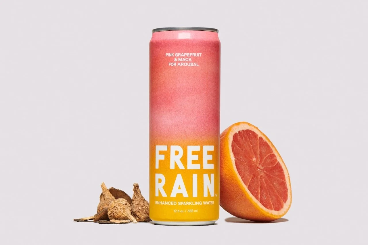 A can of sparkling water labeled 'Free Rain' with pink grapefruit and maca for arousal, placed next to half a grapefruit and some ginger root on a plain background.