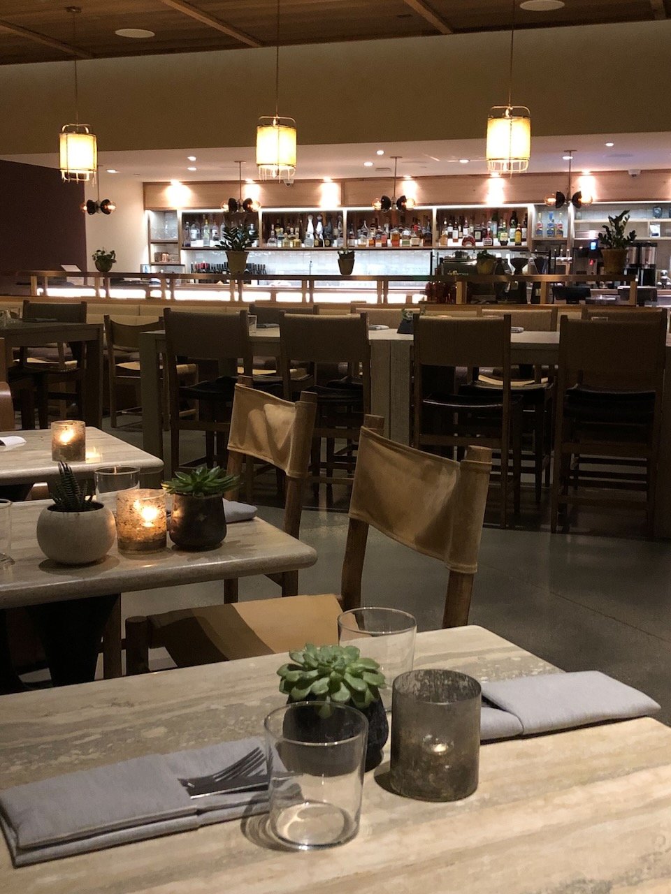 Interior of a modern restaurant with wooden furniture, candlelit tables, and a bar stocked with bottles in the background. Soft lighting creates a cozy ambiance.