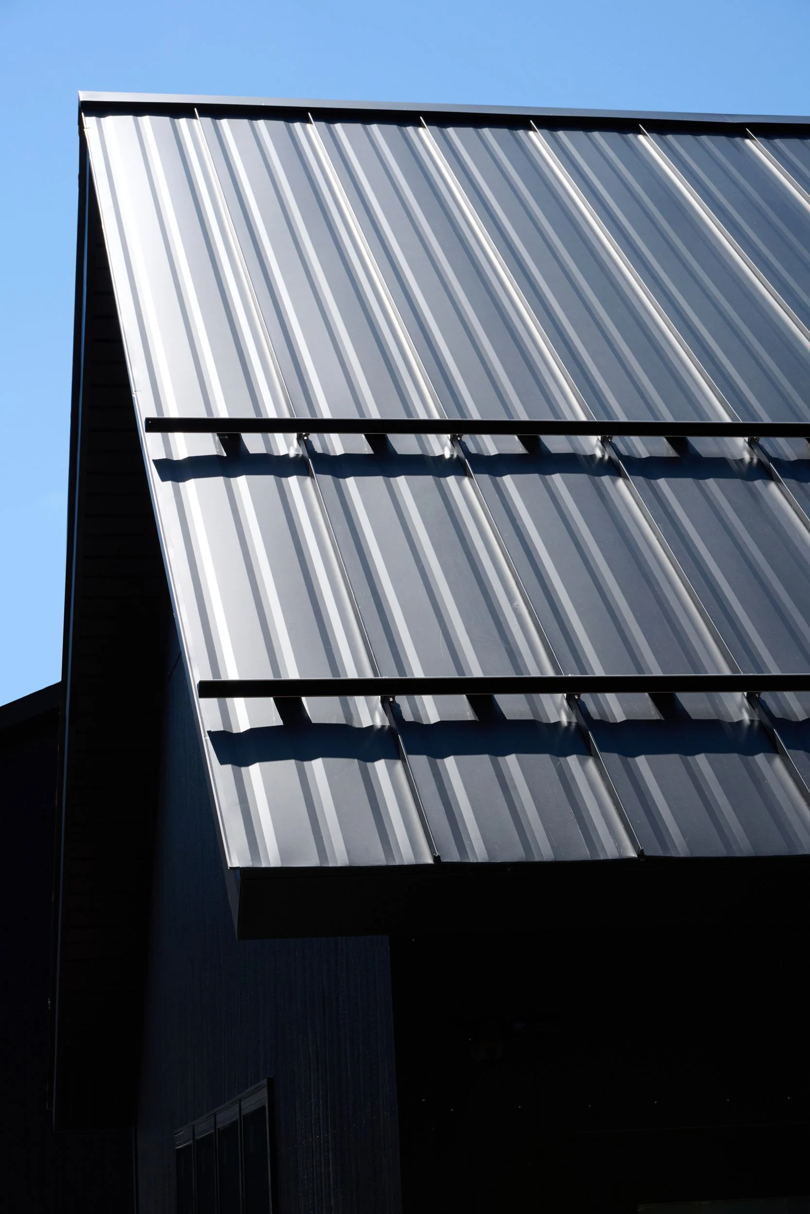 Close-up of a modern building's exterior with dark metal roofing and reflective metal panels, under a clear blue sky.
