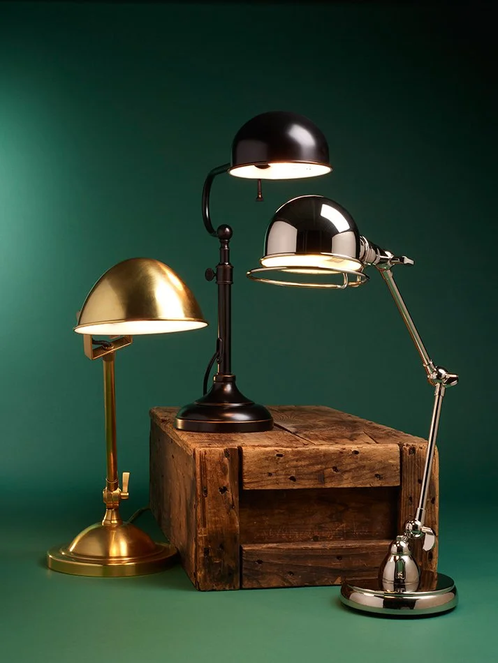 Three metallic desk lamps in black, gold, and silver, placed on a rustic wooden box against a green background.