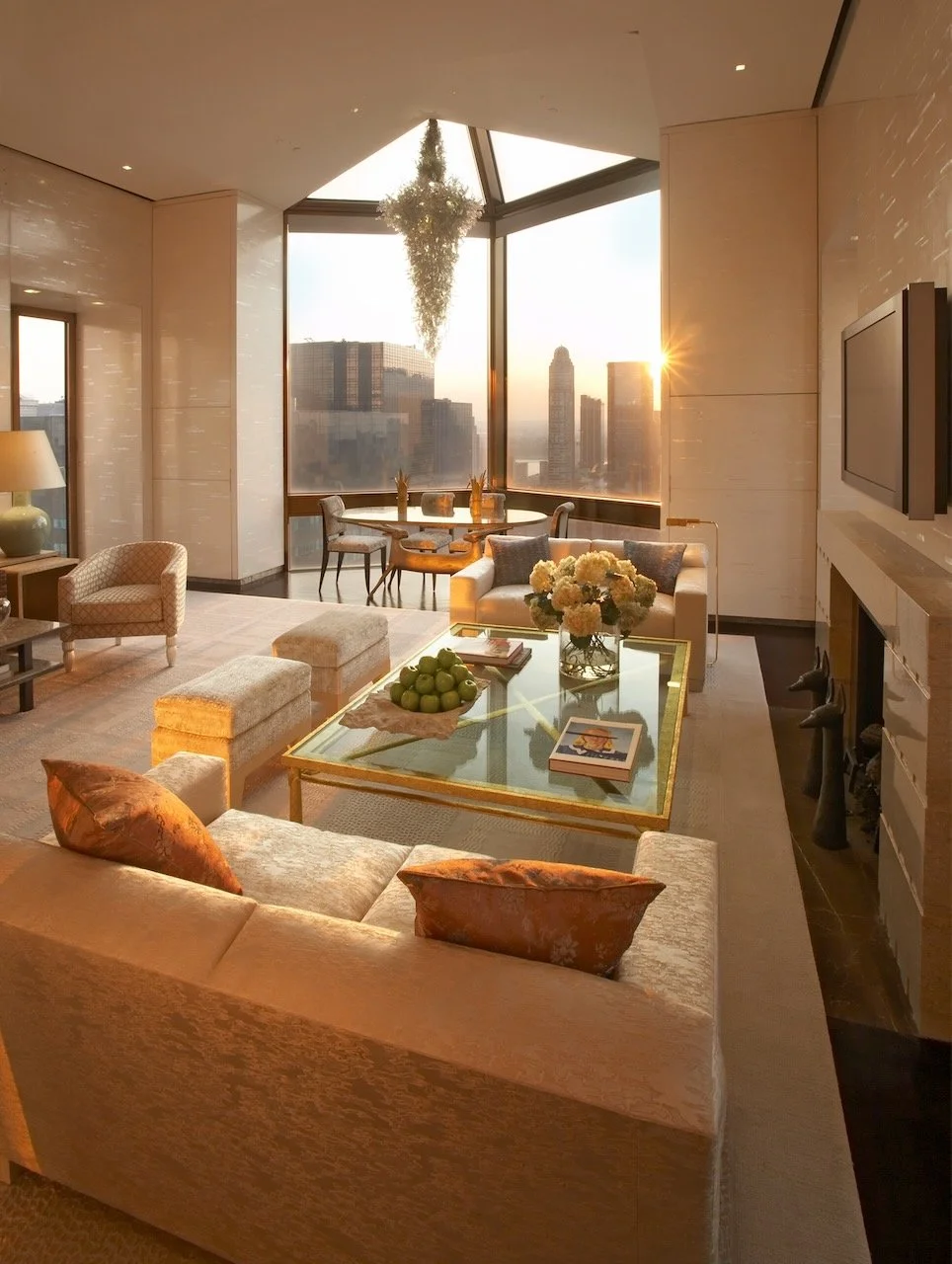 Modern living room with large windows showing city skyline at sunset, beige sofa with orange pillows, glass coffee table with flowers and green apples, a dining area with chairs, and a fireplace on the right wall.