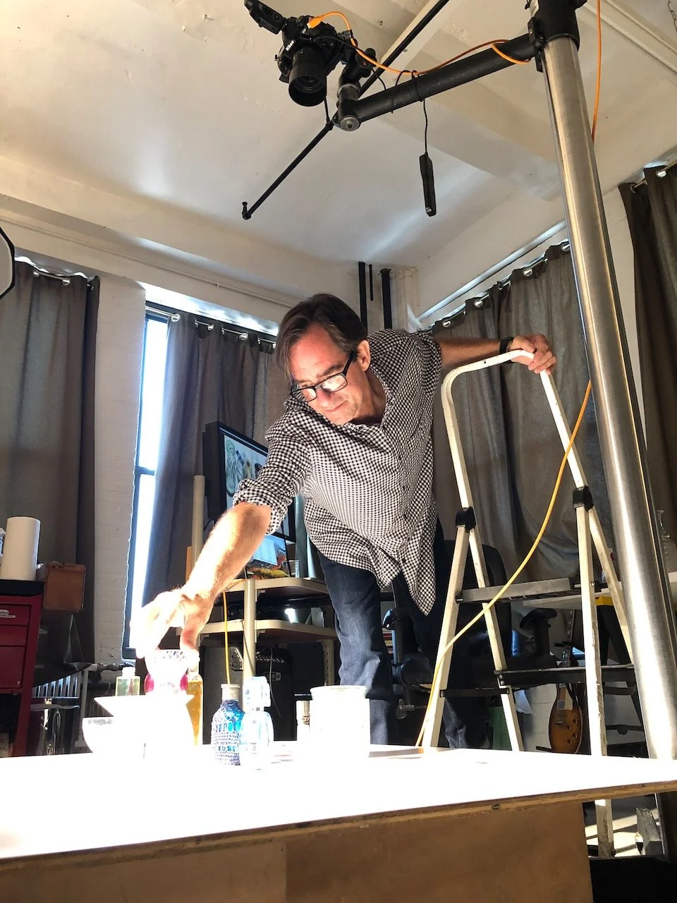 Man on a ladder reaching for items on a table in a room with cameras and dark curtains.