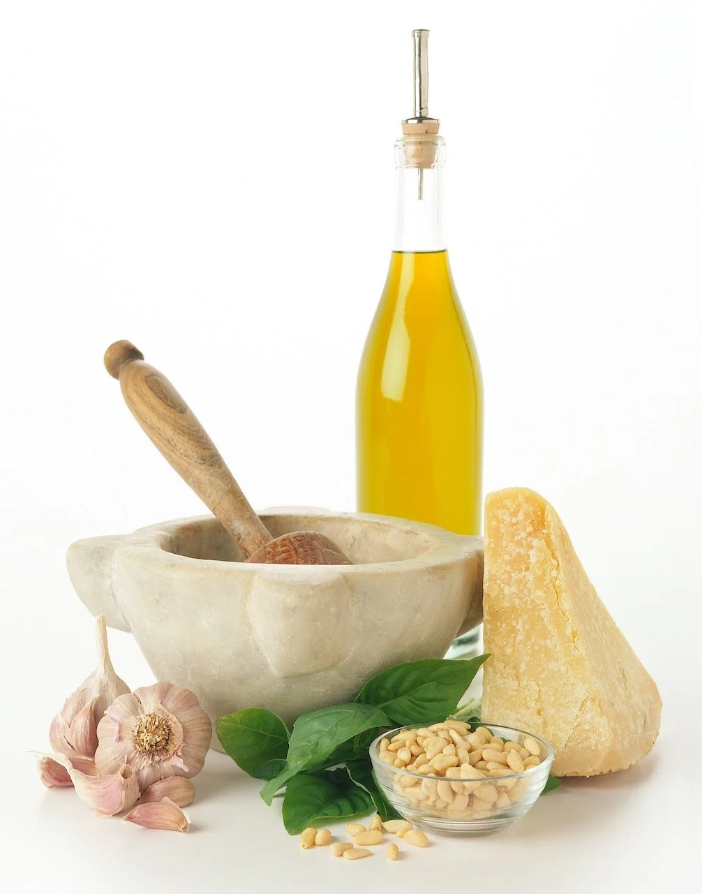 A still life of ingredients for pesto, including garlic, basil, pine nuts, olive oil, Parmesan cheese, and a pestle and mortar.