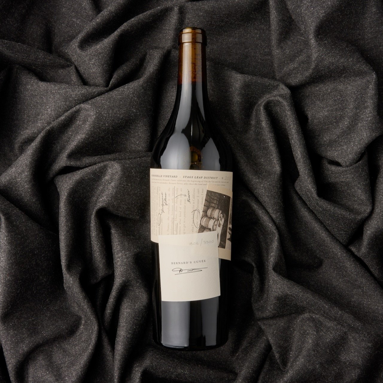 A wine bottle lying on black textured fabric, with a vintage-style label featuring handwritten text, signatures, and an image of barrels.