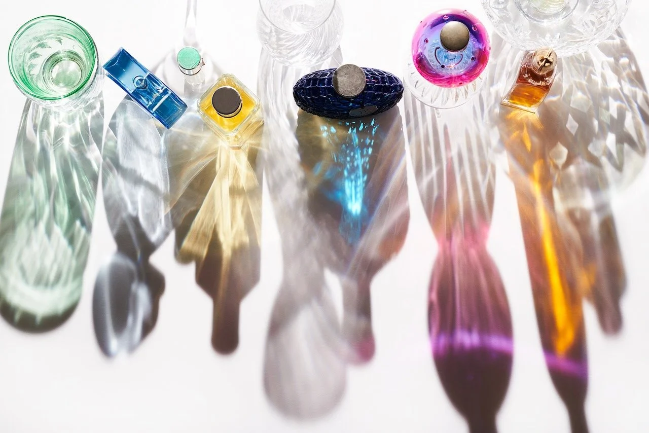 Colorful glassware pieces, including bowls and small bottles, arranged on a white surface with light creating rainbow reflections and shadows.