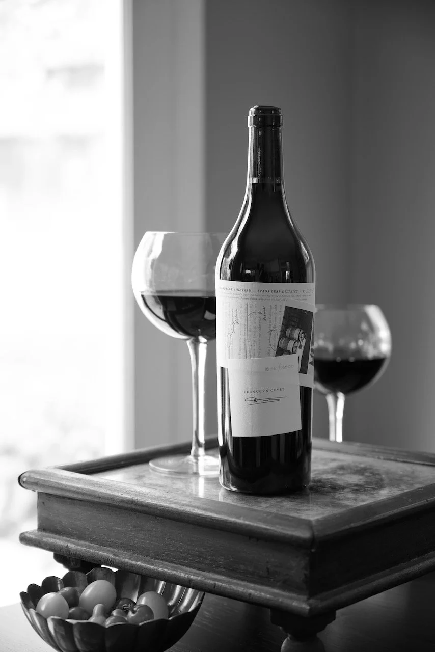 A bottle of red wine and two glasses filled with red wine placed on a wooden tray, with a bowl of eggs in front of the tray, in a black and white photo.
