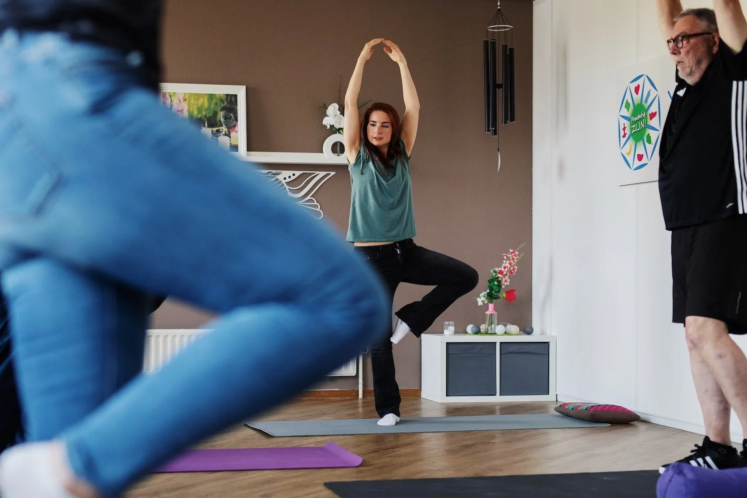 Vrouw die in een yogapose staat op een yogamat, terwijl ze haar armen omhoog strekt. Mensen die meedoen aan een indoor yogales, in een kamer met een decoratief bloemstuk en een poster met 'ZON!'.