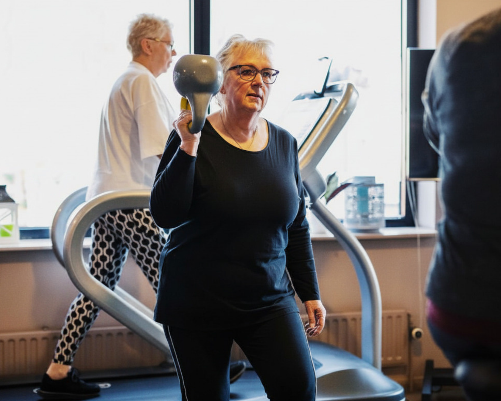 Een oudere vrouw traint in een sportschool met gewichten, terwijl een andere persoon op een loopband op de achtergrond bezig is.