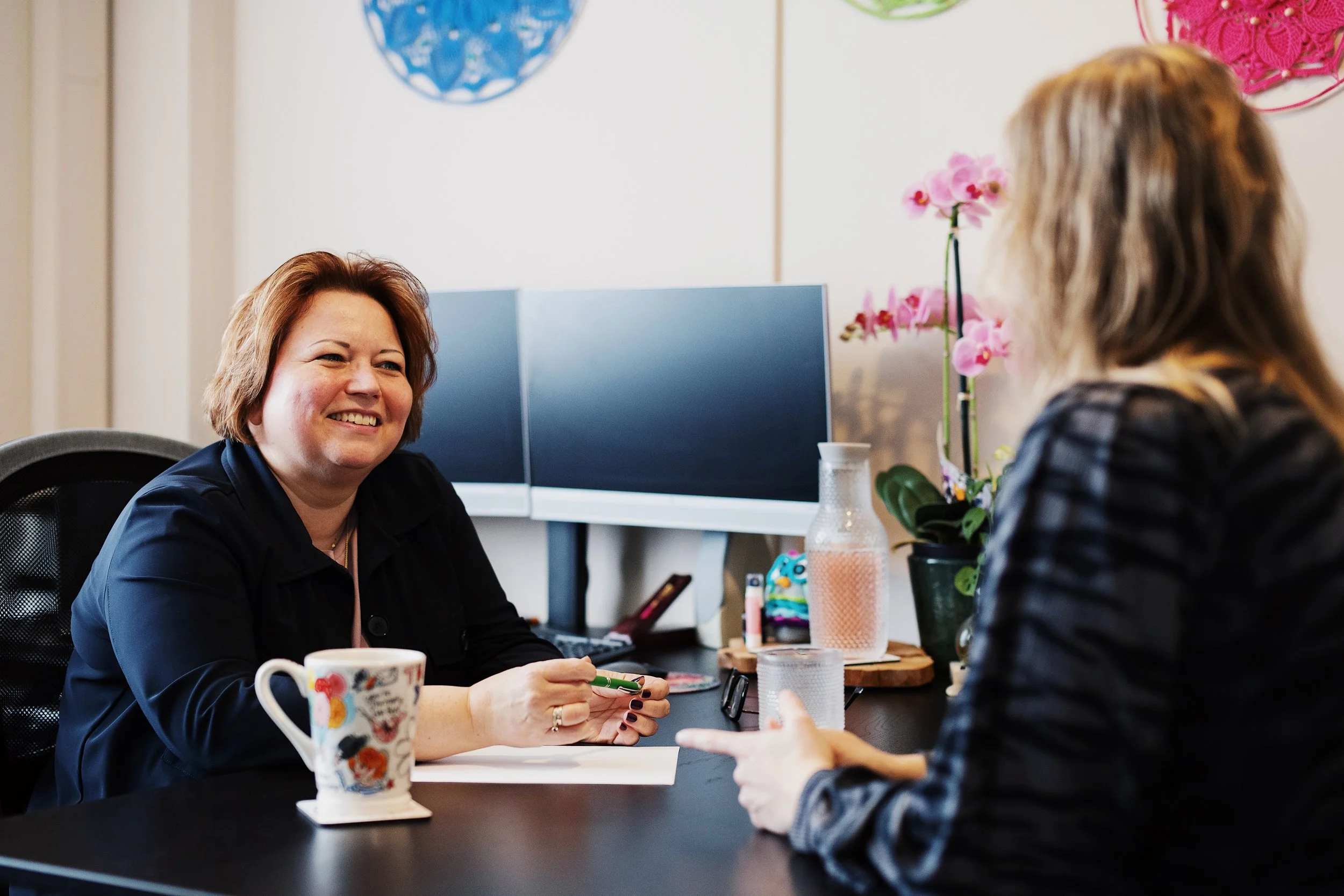 Vrouw die glimlachend aan een gesprek deelneemt, zittend aan een bureau met een vrouw die haar iets vertelt.