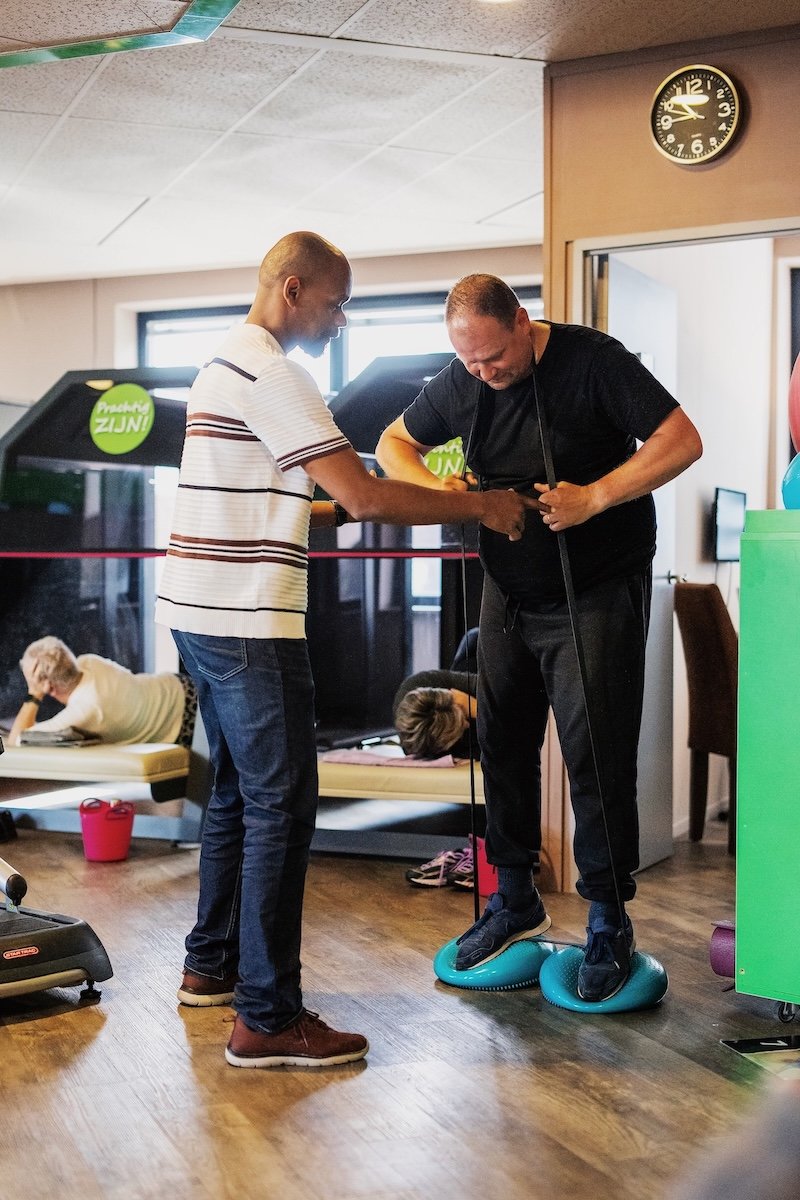 Een man op een zwart en blauwe evenwichtsbureau wordt ondersteund door een trainer van de medische fitness terwijl hij traint op een fitnessapparaat in een revalidatiecentrum, met ouderen op de achtergrond die rusten.