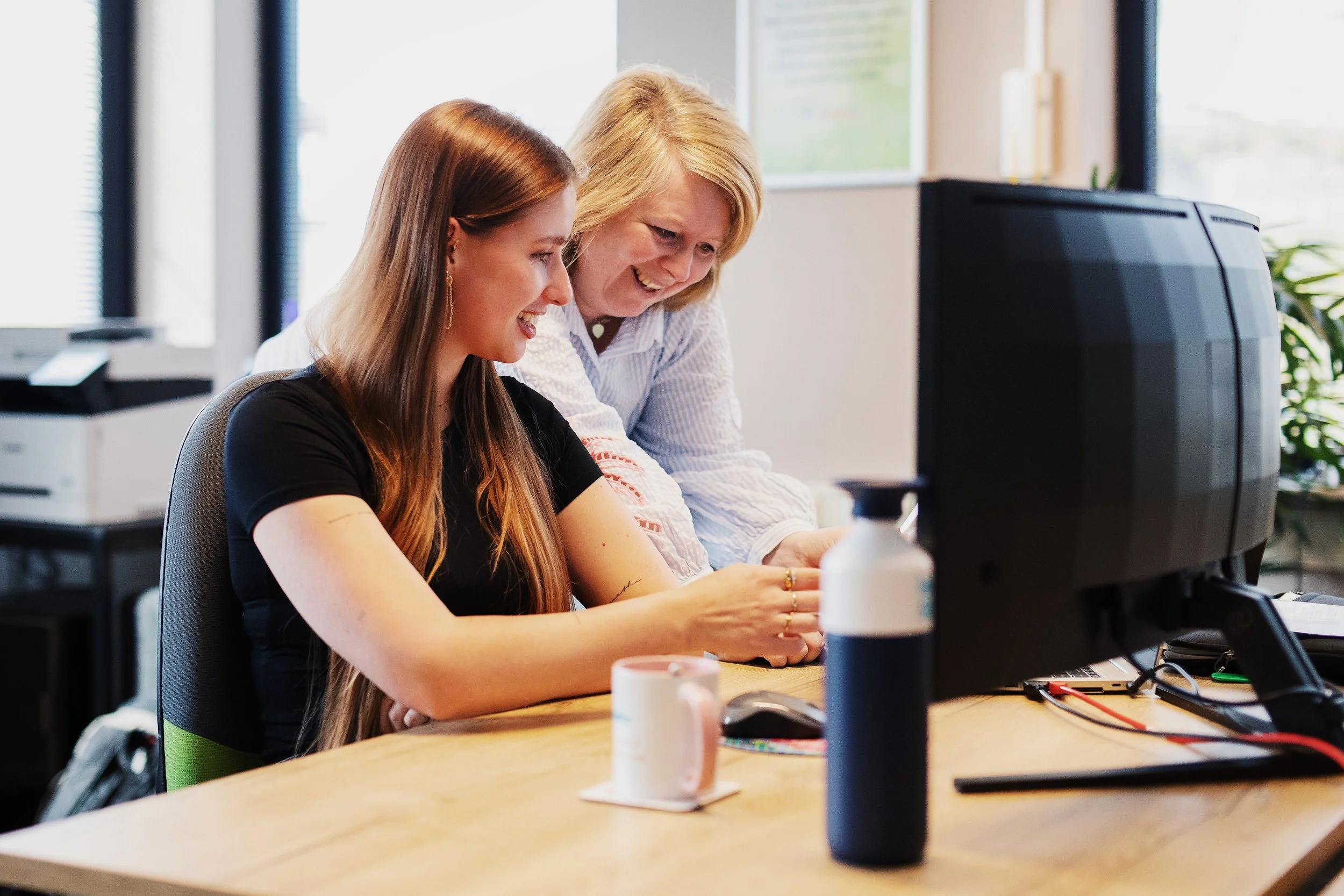 Twee vrouwen kijken samen naar een computerscherm en lachen in een kantooromgeving.