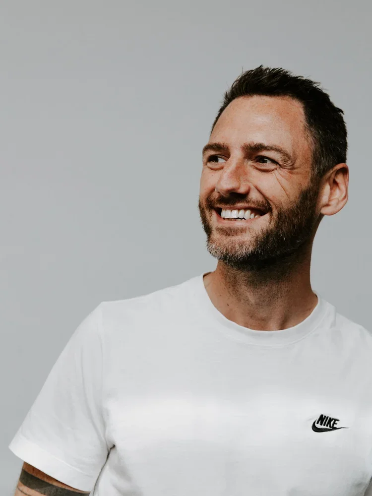 A smiling man with short dark hair and a beard, wearing a white Nike t-shirt, against a plain gray background.
