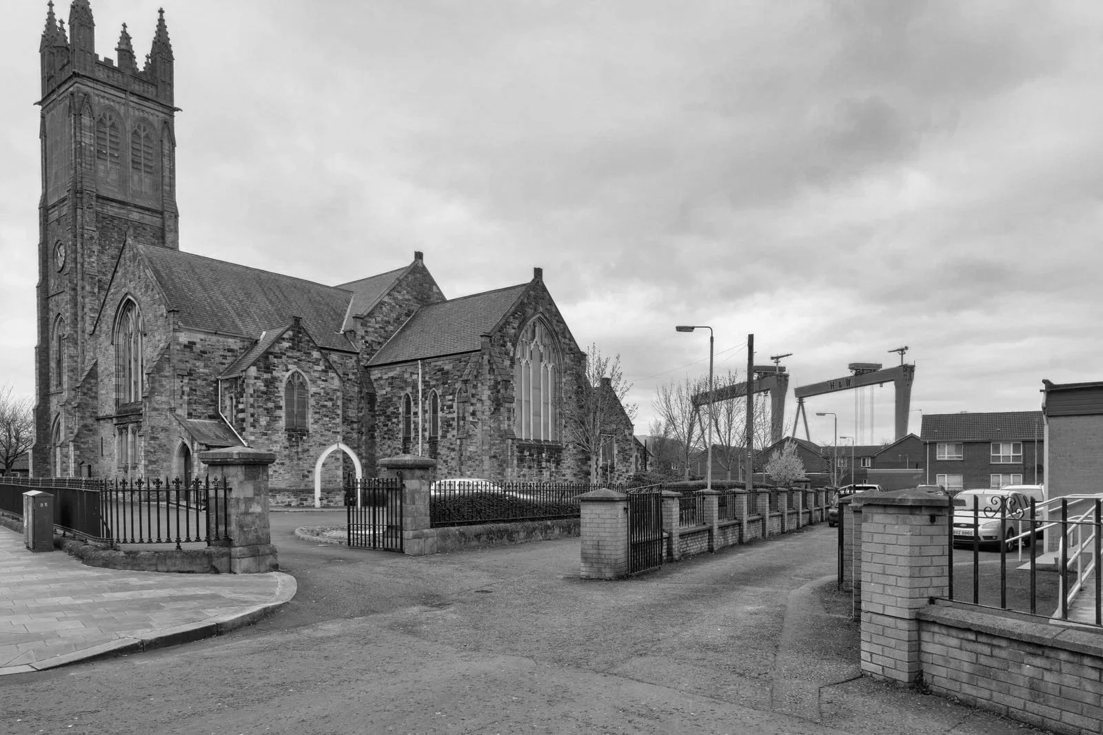 Black and white photo of a stone church with a tall clock tower, arched windows, and gated entrance, surrounded by a fence and residential houses, with overcast sky.