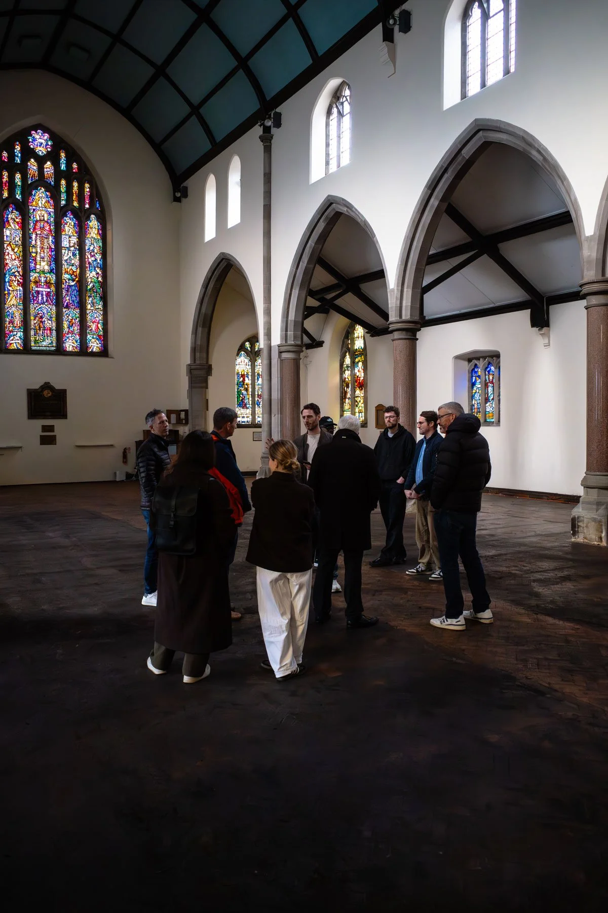Group of people standing and talking inside a church with stained glass windows, arched doorways, and wooden flooring.